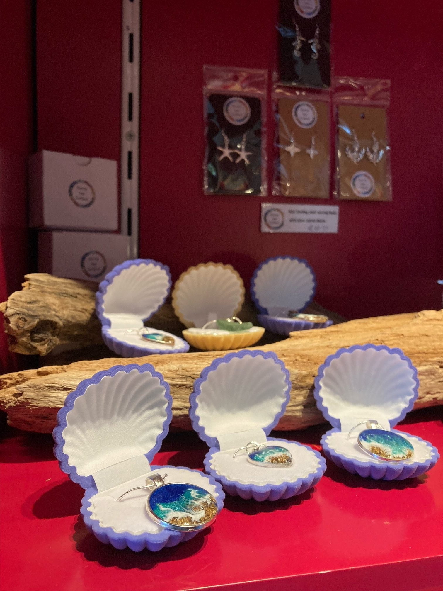 Three necklaces inside shell-shaped boxes. The necklaces have charms on them. The charms are made out of resin, and look like the sea.