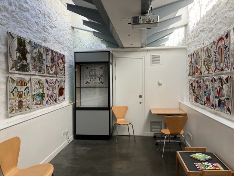 Looking in on the JMB exhibition space as if you have just entered. It is one room with textured brick walls, painted white. There are tapestries hung on the walls.